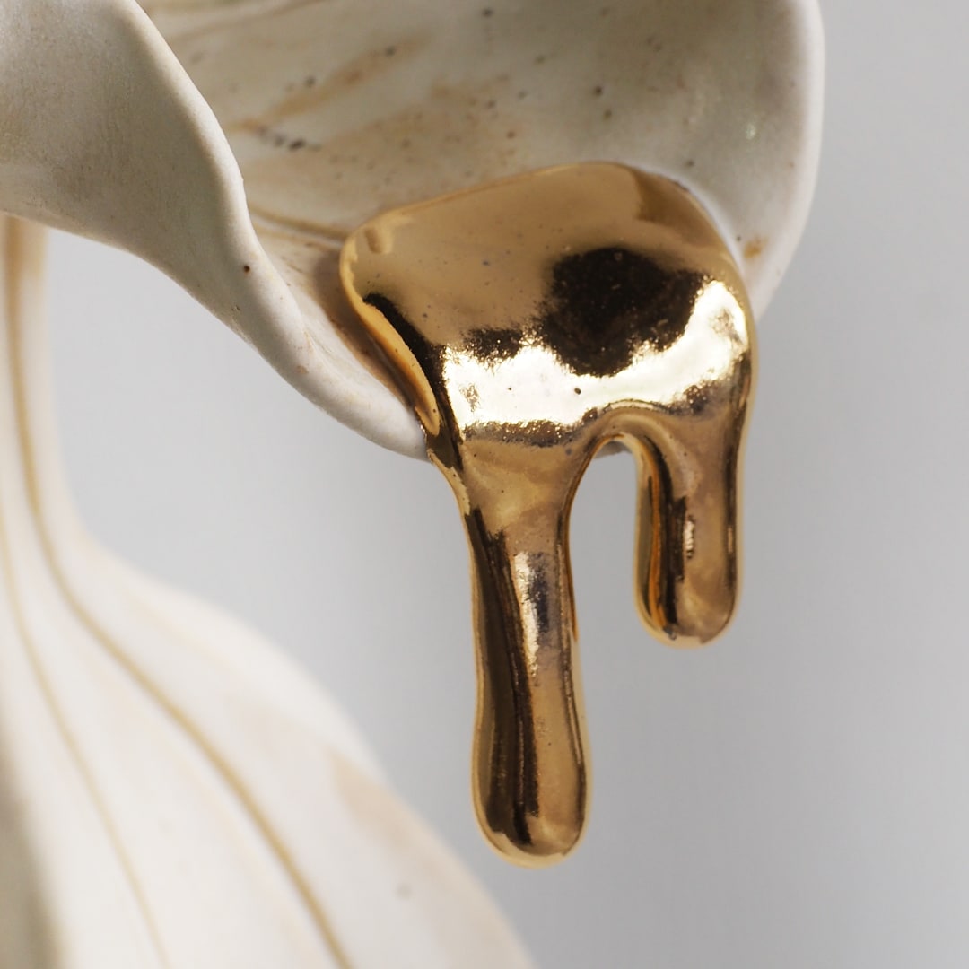 A close-up of a glossy metallic gold liquid dripping from a matte off-white ceramic container against a light gray background.