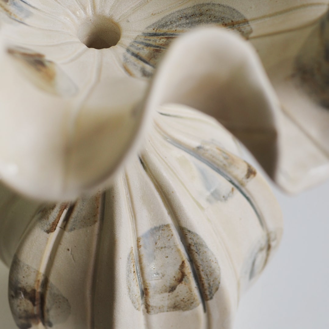 Close-up of a ceramic vase with a sculptural, flower-like form in warm off-white with gray and brown abstract painted accents.