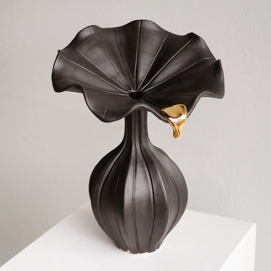 A sculptural black ceramic vase with a wide, wavy flared top resembling a lotus bloom, a ribbed bulbous base, and a small metallic gold accent on the side, displayed on a white pedestal against a light gray wall.