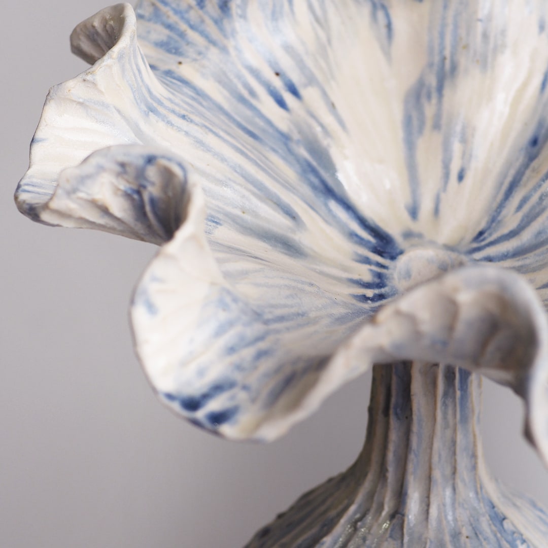 A close-up of a ceramic floral sculpture resembling a ruffled iris or flower, featuring textured off-white petals with blue and gray marbled detailing, set against a neutral gray background.