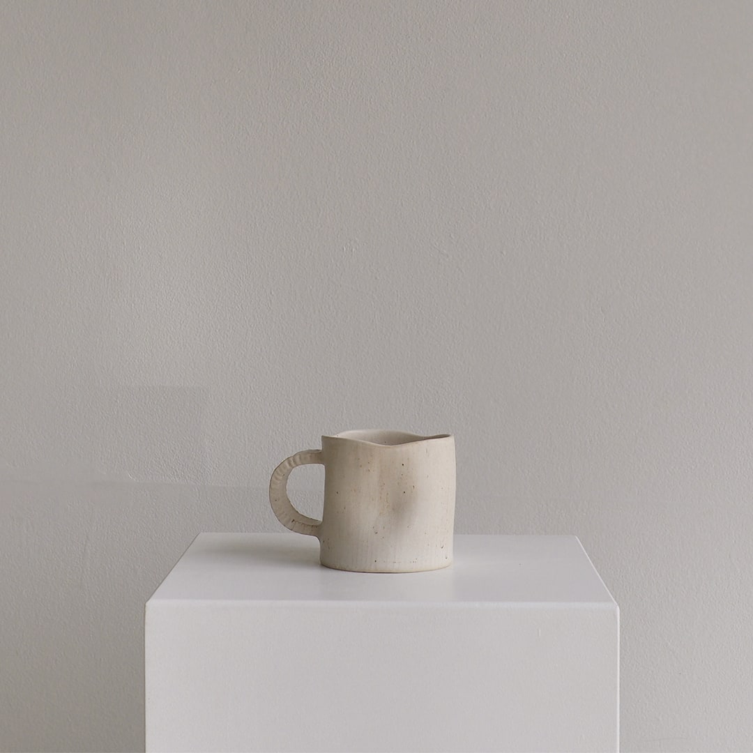 A small handmade ceramic mug with a textured, matte off-white finish and a simple loop handle, displayed on a white pedestal against a neutral gray wall.