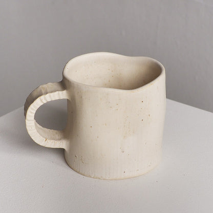 A handmade ceramic mug with a textured, organic shape and a small handle, shown on a light gray surface against a neutral gray background.