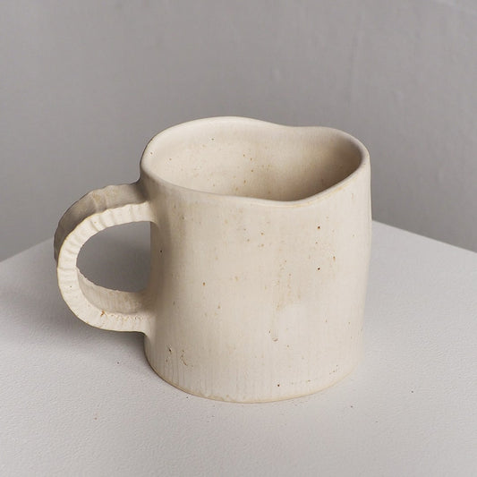 A handmade ceramic mug with a textured, organic shape and a small handle, shown on a light gray surface against a neutral gray background.