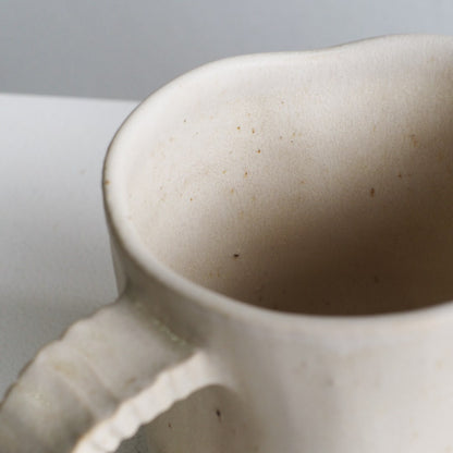 Close-up of a ceramic mug with a textured, matte off-white finish and a small handle, set against a neutral light gray background.