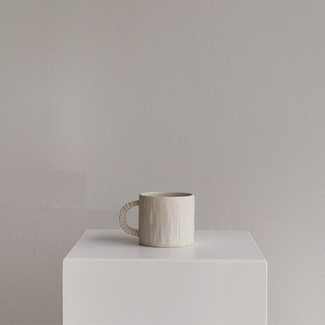 A textured ceramic mug in a warm off-white or cream color sits on a white display pedestal against a neutral gray wall.