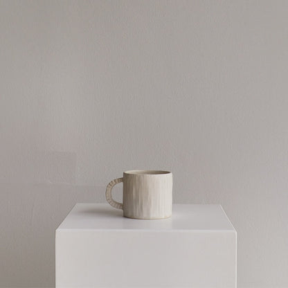 A textured ceramic mug in a warm off-white or cream color sits on a white display pedestal against a neutral gray wall.