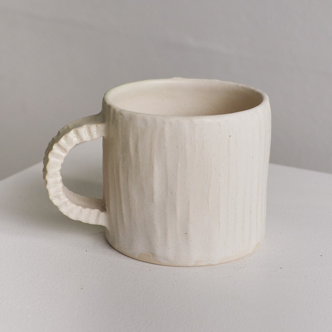An off-white ceramic mug with a vertically ribbed textured surface and a fluted handle, shown on a neutral light gray surface.