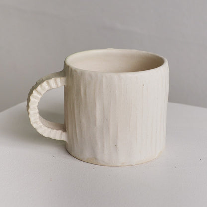 An off-white ceramic mug with a vertically ribbed textured surface and a fluted handle, shown on a neutral light gray surface.