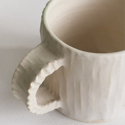 A close-up of a textured ceramic mug with a flared handle and a matte off-white finish, set against a plain light background.