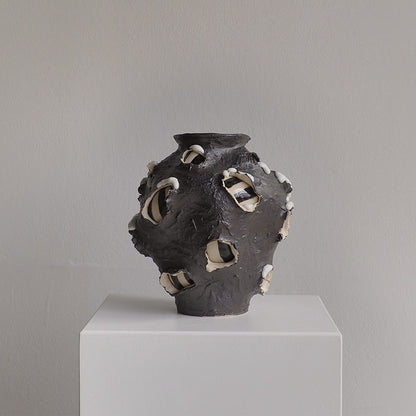 A sculptural ceramic vase with a dark charcoal body and irregular white and black glazed openings, displayed on a white pedestal against a light gray wall.