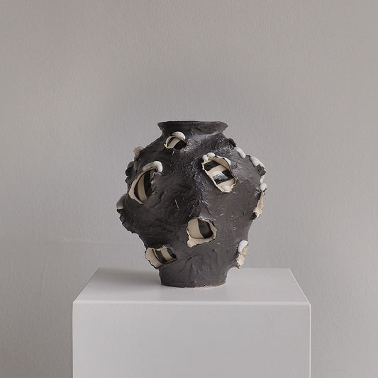 A sculptural ceramic vase with a dark charcoal body and irregular white and black glazed openings, displayed on a white pedestal against a light gray wall.