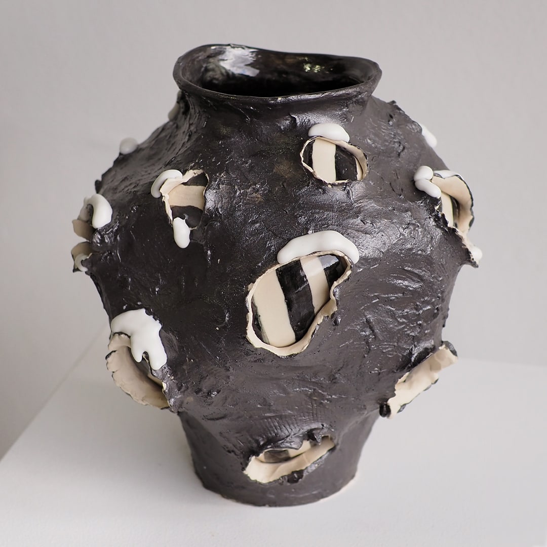 A dark charcoal-brown ceramic vase with a textured, organic surface and irregular white and cream protruding petal-like or shell-like accents, photographed on a neutral light gray background.