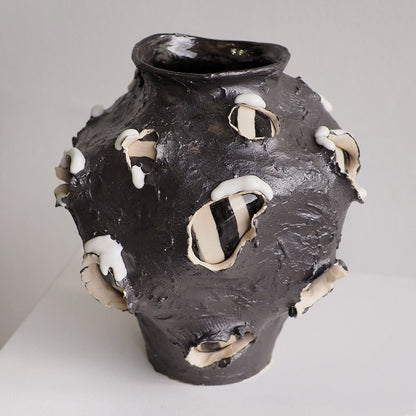 A dark charcoal-brown ceramic vase with a textured, organic surface and irregular white and cream protruding petal-like or shell-like accents, photographed on a neutral light gray background.