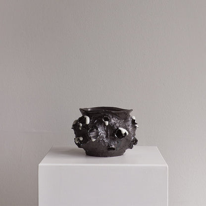A dark, textured ceramic pot with irregular raised bumps and white speckled accents, displayed on a white pedestal against a neutral gray wall.