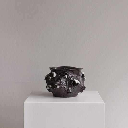 A dark, textured ceramic pot with irregular raised bumps and white speckled accents, displayed on a white pedestal against a neutral gray wall.