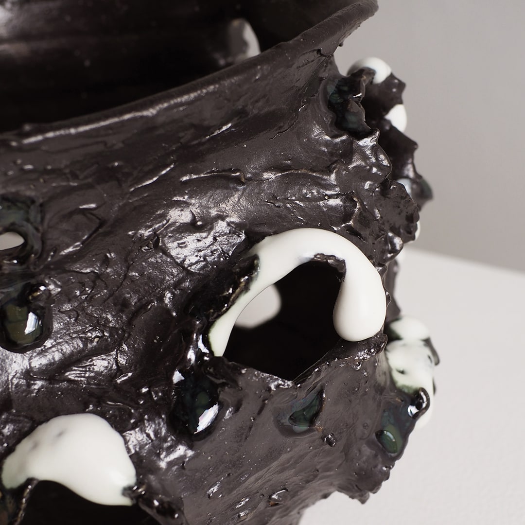 A close-up of a sculptural black vessel or planter with glossy, textured surfaces and irregular white drips, photographed against a light neutral background.