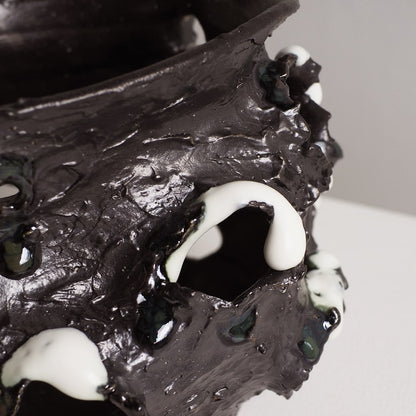A close-up of a sculptural black vessel or planter with glossy, textured surfaces and irregular white drips, photographed against a light neutral background.