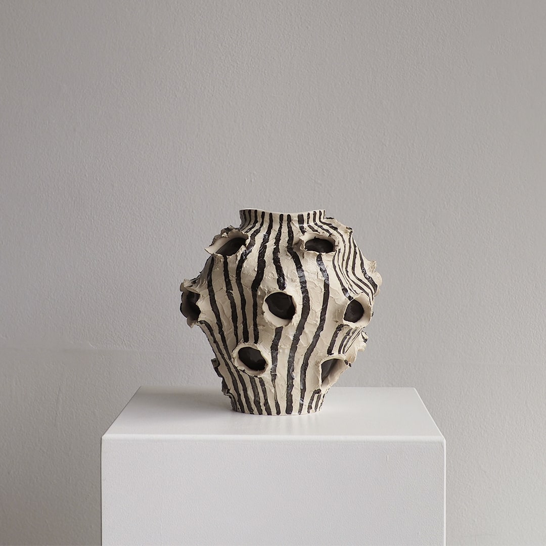 A sculptural ceramic vase with a black and off-white zebra-like vertical stripe pattern and multiple organic cutouts, displayed on a white pedestal against a neutral gray wall.