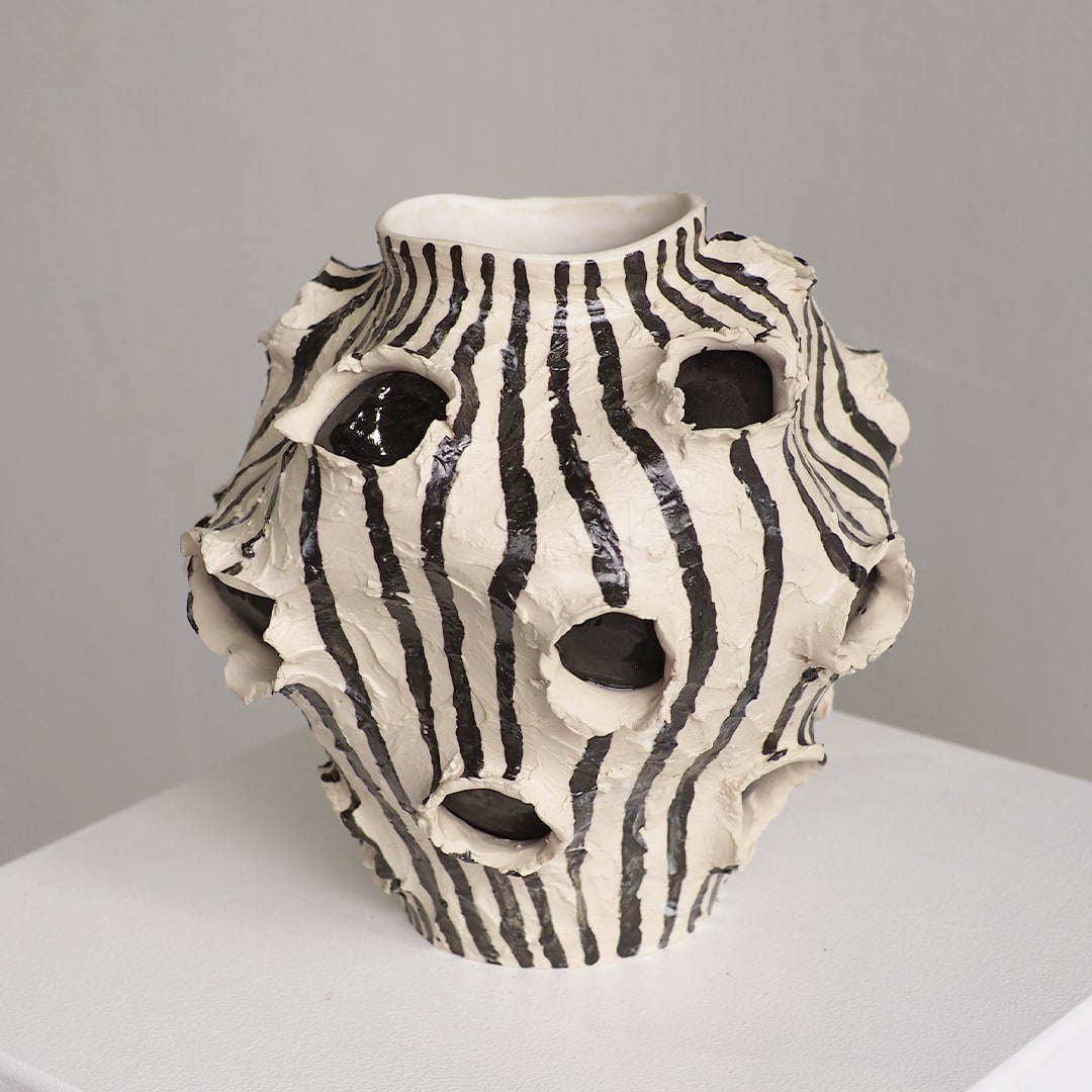 An organic-shaped ceramic vase with a zebra-like black and off-white striped pattern and multiple irregular cutouts, displayed on a white surface against a neutral gray background.