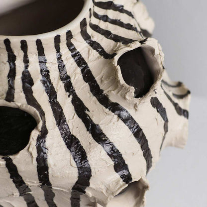A close-up of a ceramic vessel with a zebra-stripe pattern in black and off-white, featuring a large circular opening at the top and textured, hand-painted details.