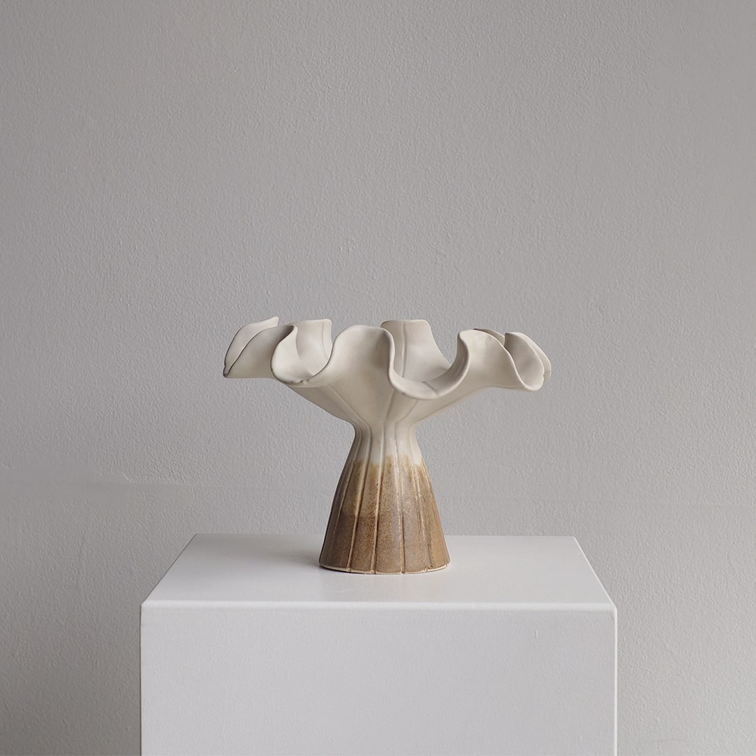 A sculptural ceramic vase with a flared, ruffled rim and a fluted pedestal base, displayed on a white plinth against a light gray wall.