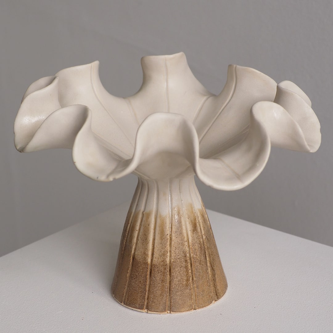 A sculptural ceramic vase with a flared, ruffled top and a fluted pedestal base, shown on a white surface against a light gray wall. The vase is off-white at the top and transitions to a warm tan glazed finish at the bottom.