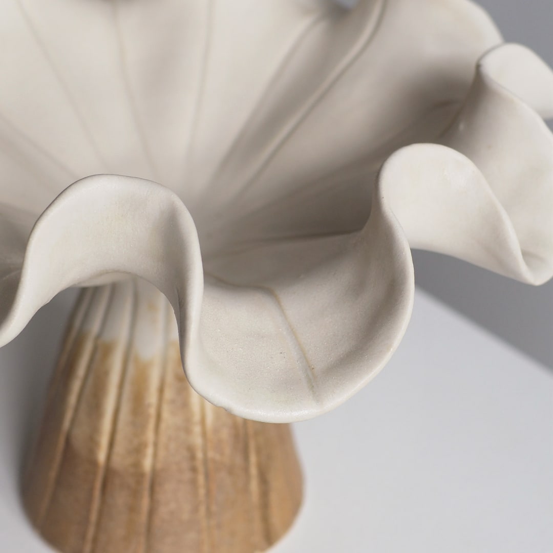 Close-up of a ceramic flower vase with ruffled petals in warm off-white and a fluted base in beige and light brown tones.