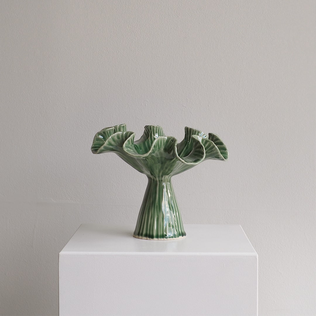 A green glazed ceramic vase with a fluted, ruffled rim and a ribbed pedestal base, displayed on a white plinth against a light gray wall.