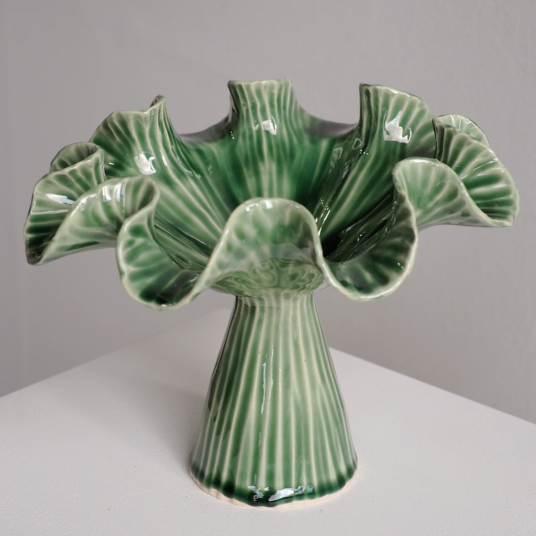 A green glazed ceramic pedestal flower pot with ruffled petals and vertical ribbed detailing, shown on a white surface against a gray background.