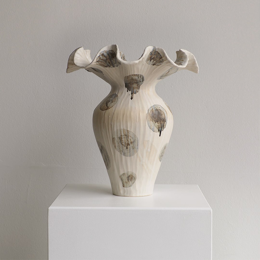 A sculptural ceramic vase with a flared, ruffled rim and ribbed body, shown on a white pedestal against a light gray wall. The vase is off-white with irregular gray and brown abstract circular marks and dripped accents.