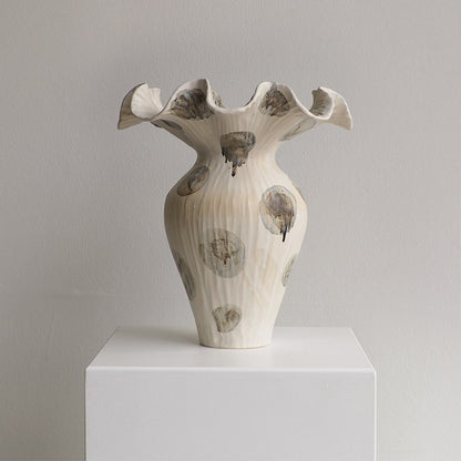 A sculptural ceramic vase with a flared, ruffled rim and ribbed body, shown on a white pedestal against a light gray wall. The vase is off-white with irregular gray and brown abstract circular marks and dripped accents.