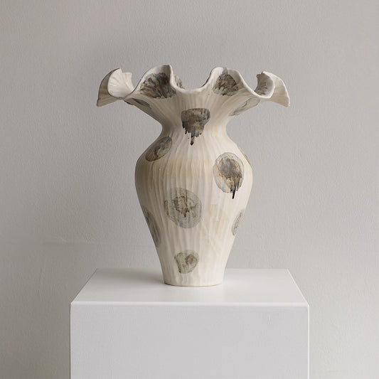 A sculptural ceramic vase with a flared, ruffled rim and ribbed body, shown on a white pedestal against a light gray wall. The vase is off-white with irregular gray and brown abstract circular marks and dripped accents.