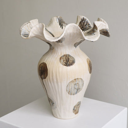 A sculptural ceramic vase with a flared, ruffled rim and ribbed body, shown on a white pedestal against a light gray wall. The vase is predominantly off-white/cream with irregular brown and gray spotted accents.
