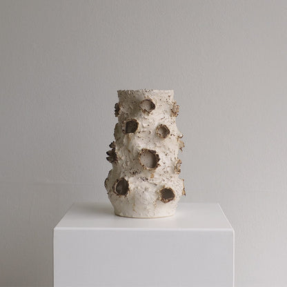 A sculptural ceramic vase with a textured, organic surface and multiple irregular holes, displayed on a white pedestal against a neutral gray background.