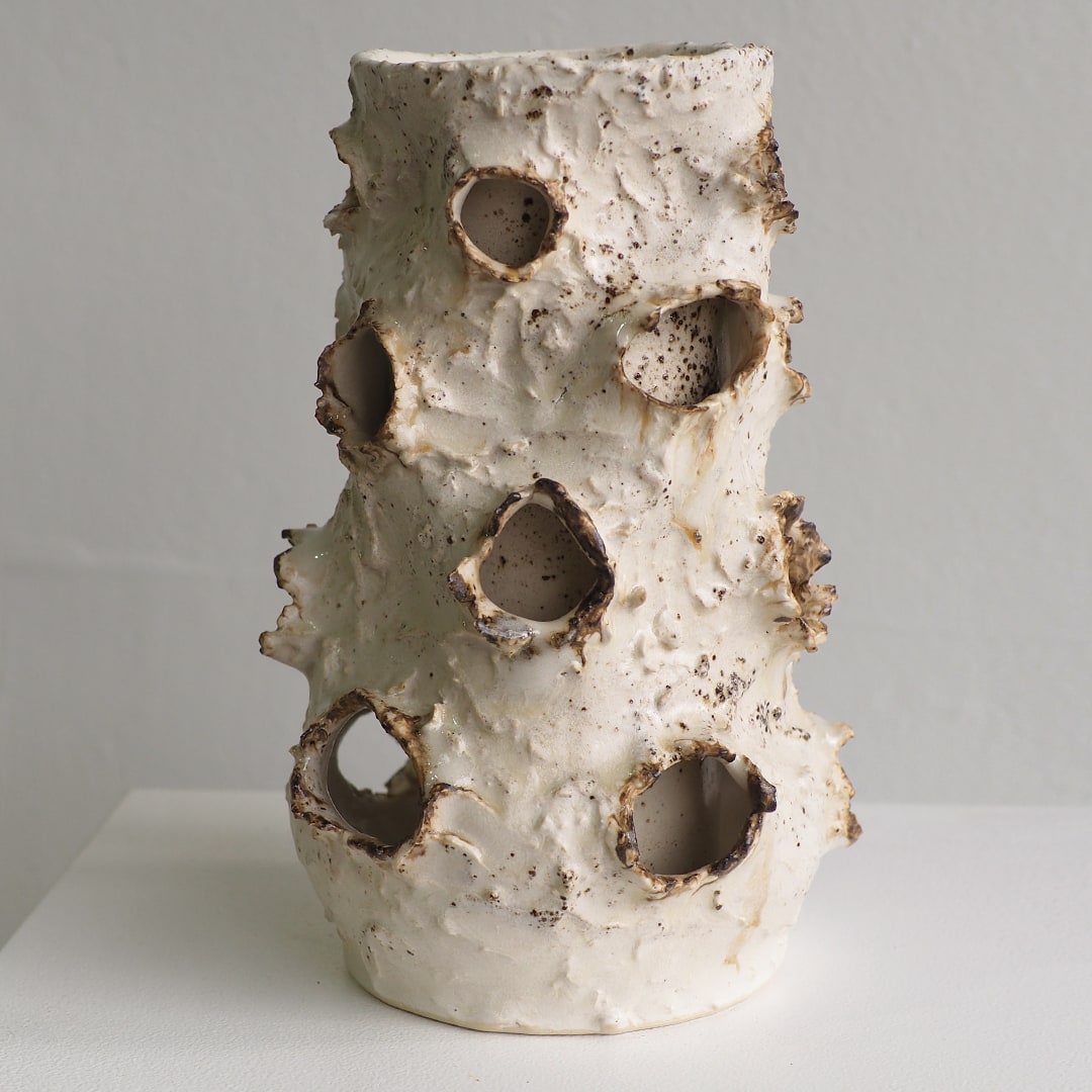 A sculptural ceramic vase with a textured, organic shape and multiple irregular hollow holes, shown on a white surface against a light gray background.