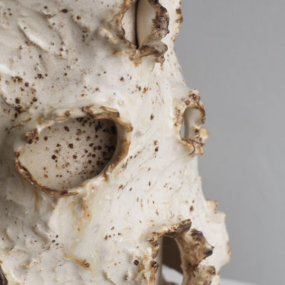 Close-up of a textured ceramic sculpture with a speckled off-white glaze and dark brown details, set against a neutral gray background.