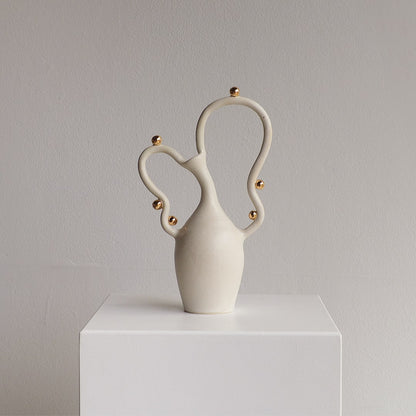 A sculptural ceramic vase in matte off-white with small metallic gold accents, displayed on a white pedestal against a light gray wall.