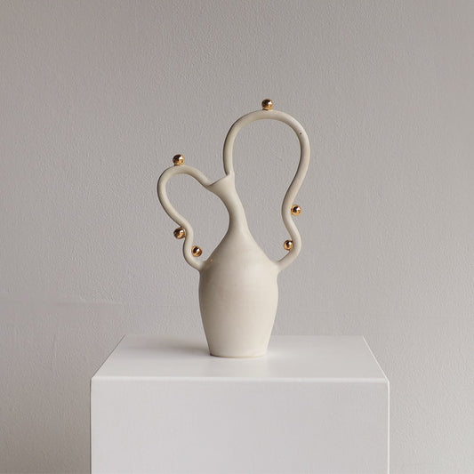 A sculptural ceramic vase in matte off-white with small metallic gold accents, displayed on a white pedestal against a light gray wall.