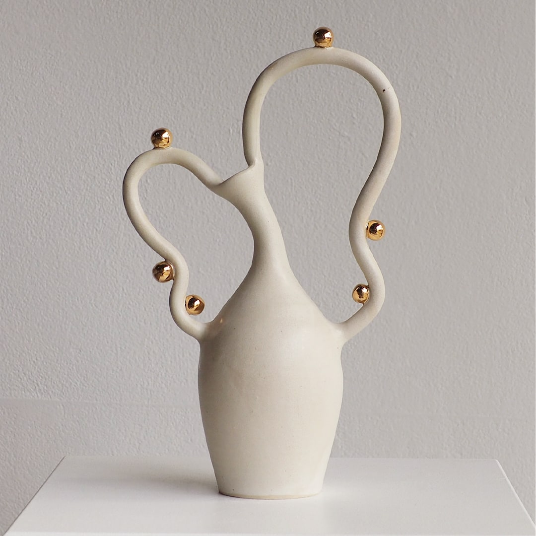 An off-white ceramic vase with a unique sculptural design, featuring two curved handles adorned with small gold metallic beads, set against a neutral light gray background.