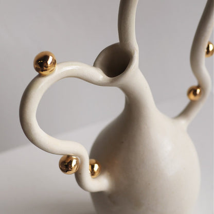 A close-up of a modern ceramic vase with a matte off-white body and shiny gold metallic accents on its curved handle and neck.