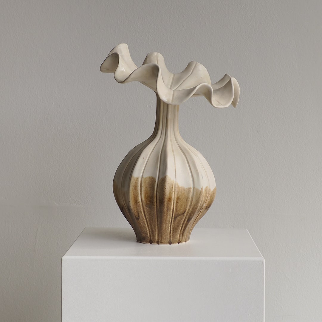 An abstract ceramic vase with a flared, wavy top and ribbed bulbous body, shown on a white display pedestal against a light gray wall.