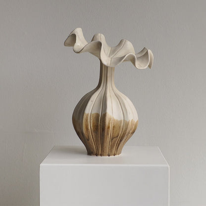 An abstract ceramic vase with a flared, wavy top and ribbed bulbous body, shown on a white display pedestal against a light gray wall.