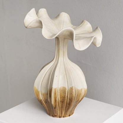 A sculptural ceramic vase with a flared, wavy top and ribbed bulbous base, shown on a white surface against a light gray wall. The vase is off-white/cream with a warm brown glazed lower section.