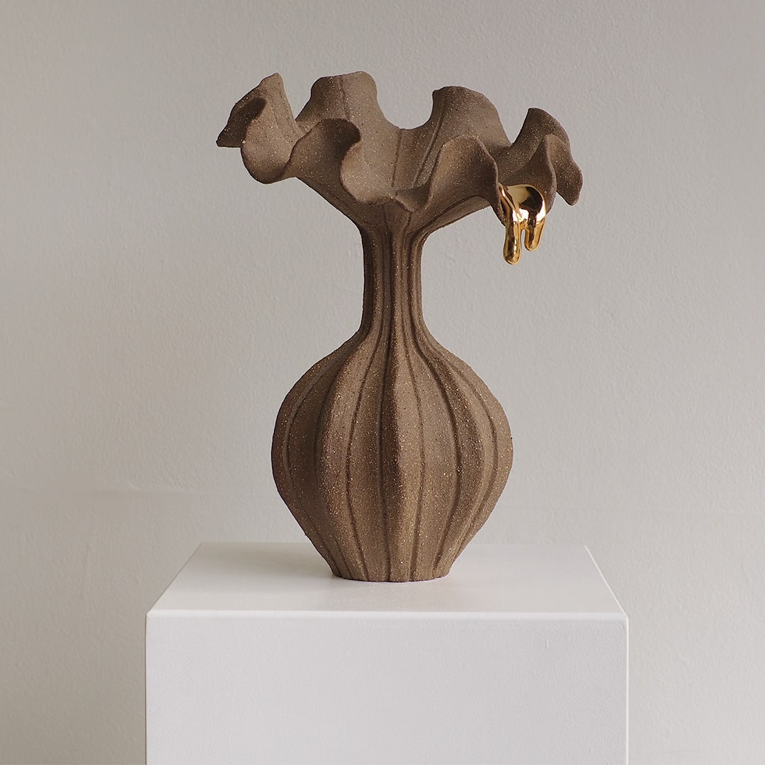 A sculptural ceramic vase with a ribbed bulbous base and flared, wavy rim, shown in a warm taupe-brown glaze with a small gold accent at the top, displayed on a white pedestal against a light gray wall.
