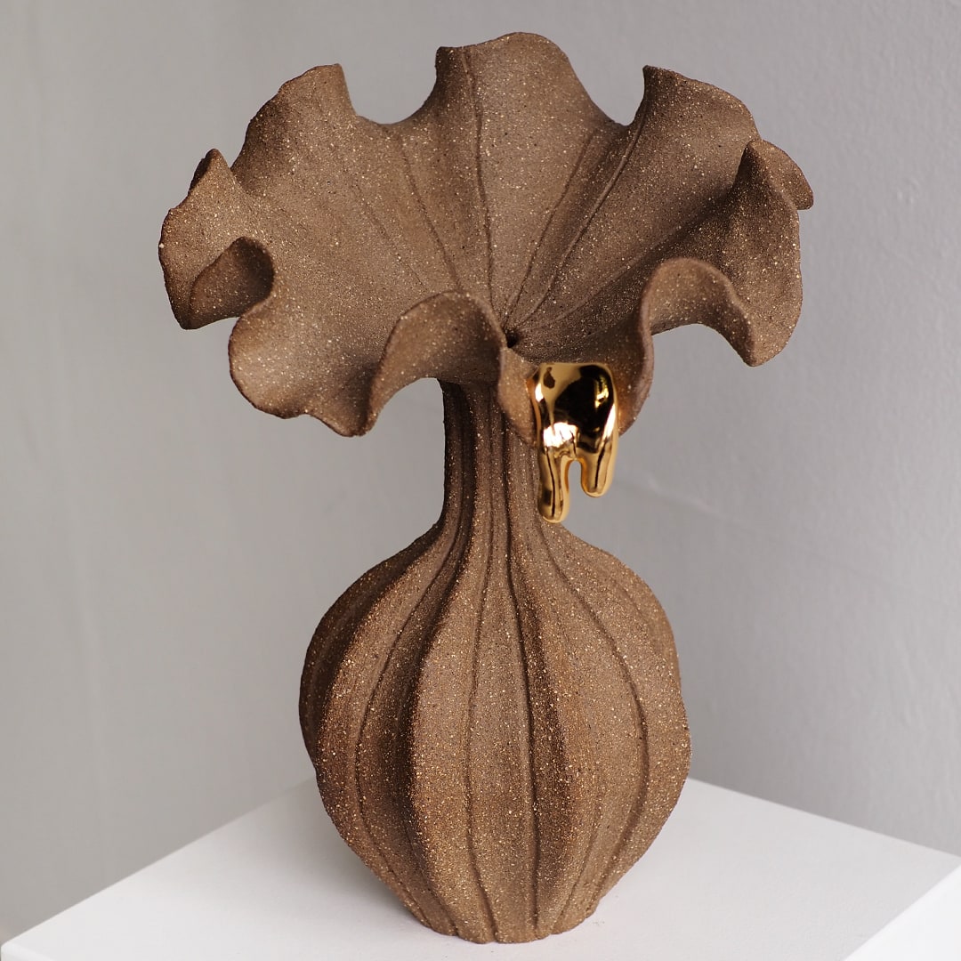 A sculptural brown vase with a textured, ribbed bulbous base and a wide, ruffled top, accented by a small dripping gold element near the rim, set against a neutral gray background.