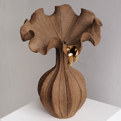 A sculptural brown vase with a textured, ribbed bulbous base and a wide, ruffled top, accented by a small dripping gold element near the rim, set against a neutral gray background.