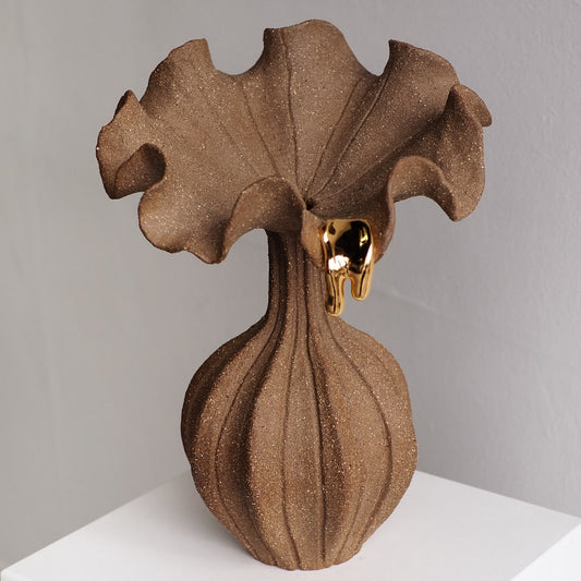 A sculptural brown vase with a textured, ribbed bulbous base and a wide, ruffled top, accented by a small dripping gold element near the rim, set against a neutral gray background.