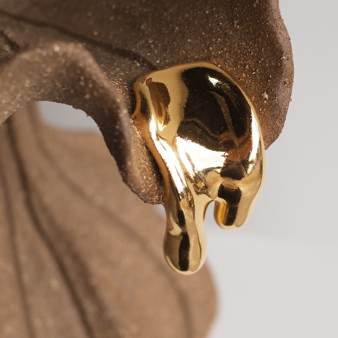 A close-up of a glossy, dripping gold earring or pendant resting against a textured brown surface, with a soft light gray background.