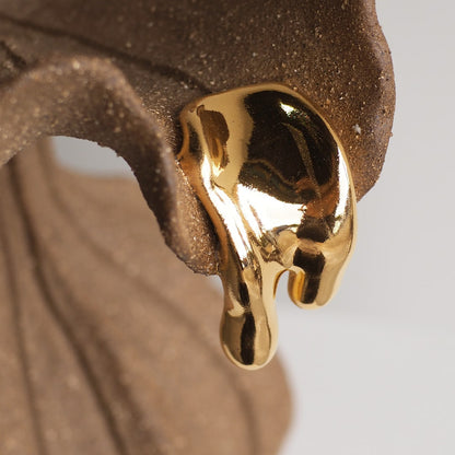 A close-up of a glossy, dripping gold earring or pendant resting against a textured brown surface, with a soft light gray background.