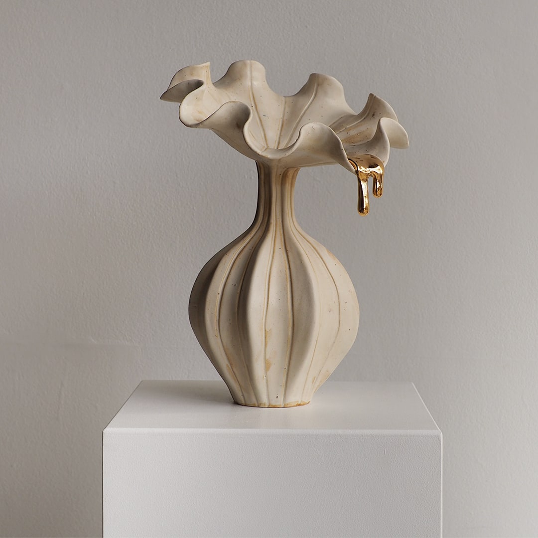 A sculptural ceramic vase with a flared, ruffled top and ribbed bulbous body, shown in a warm off-white/cream tone on a white pedestal against a light gray wall.